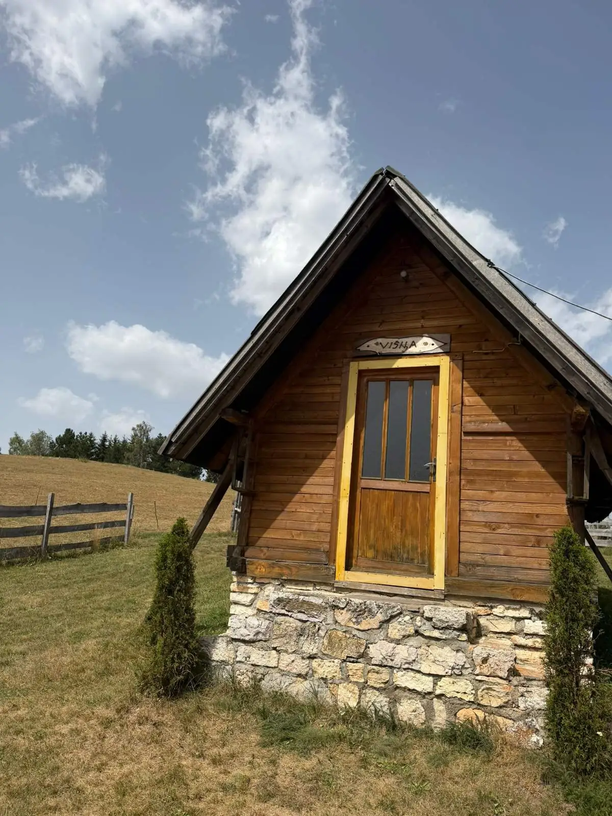 Mala drvena vikendica na kamenom postolju, sa žutim ramom vrata i natpisom Višnja